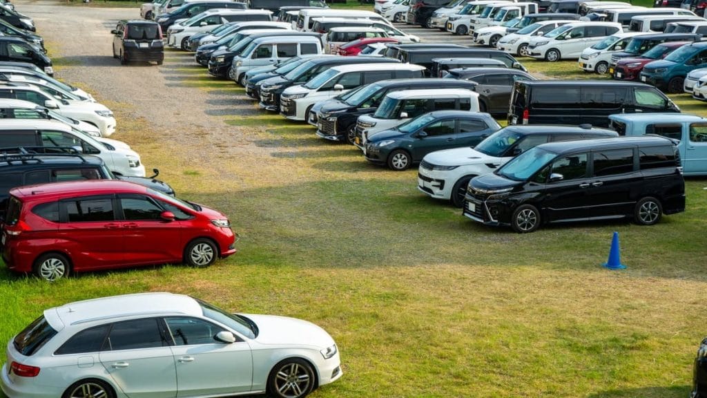 芝生と砂利の屋外駐車場に駐車された車の列。白、黒、カラーの車が混在し、青い交通コーンが見える。